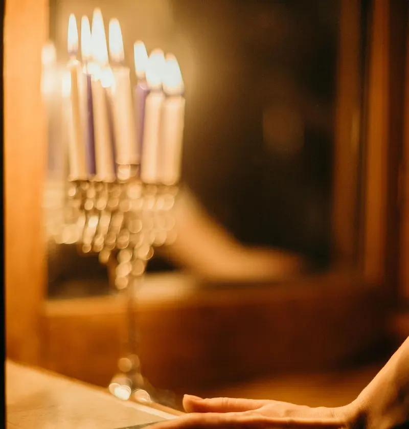 A menorah's candles burning out of focus in warm light, with a hand resting on the table in front.
